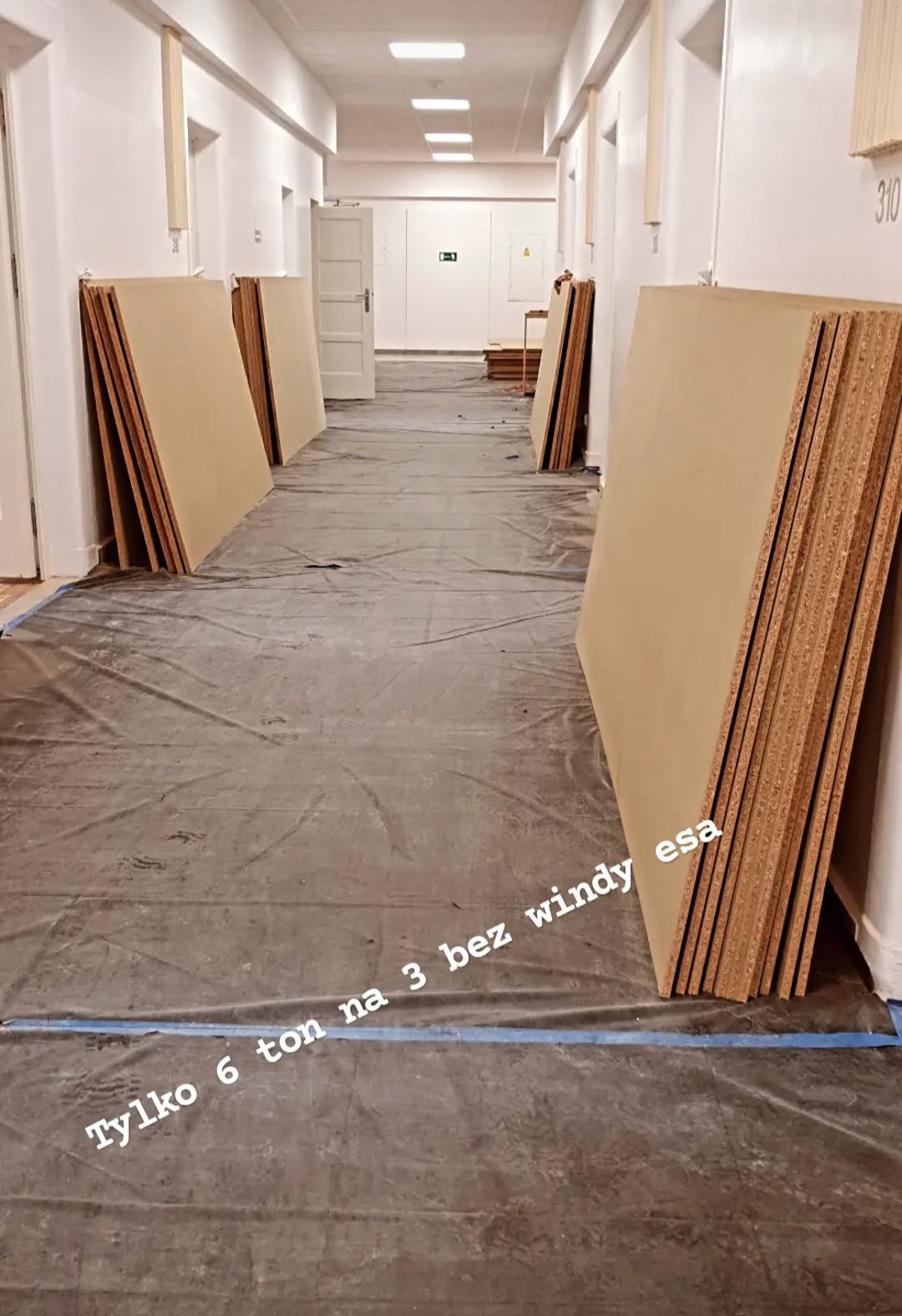 Korytarz zabezpieczony folią malarską, z ułożonymi płytami MDF pod ścianami. Widoczne oznaczenia na ścianie i tekst na folii. Przygotowanie do prac wykończeniowych.