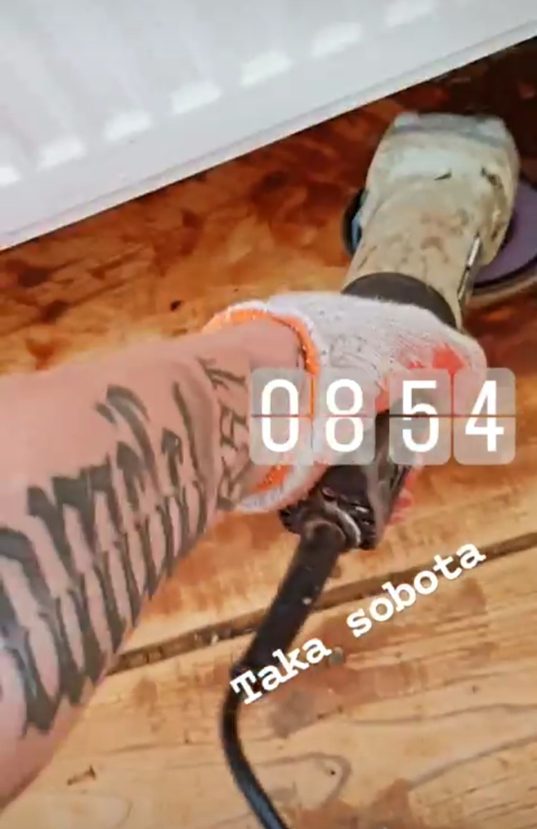 Szlifowanie drewnianej podłogi szlifierką oscylacyjną przez osobę z tatuażem na ramieniu, w rękawicy roboczej. Widoczne deski podłogowe i fragment listwy przypodłogowej.