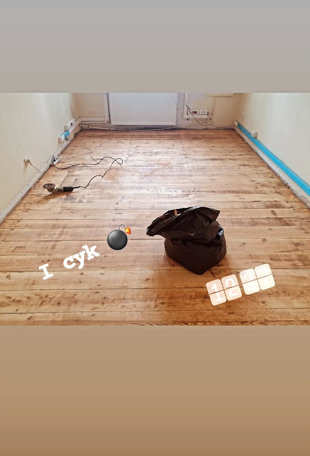 Pokój z drewnianą podłogą w trakcie renowacji. Widoczna szlifierka kątowa, worek na śmieci i zabawne naklejki. Podłoga wymaga dalszej obróbki.