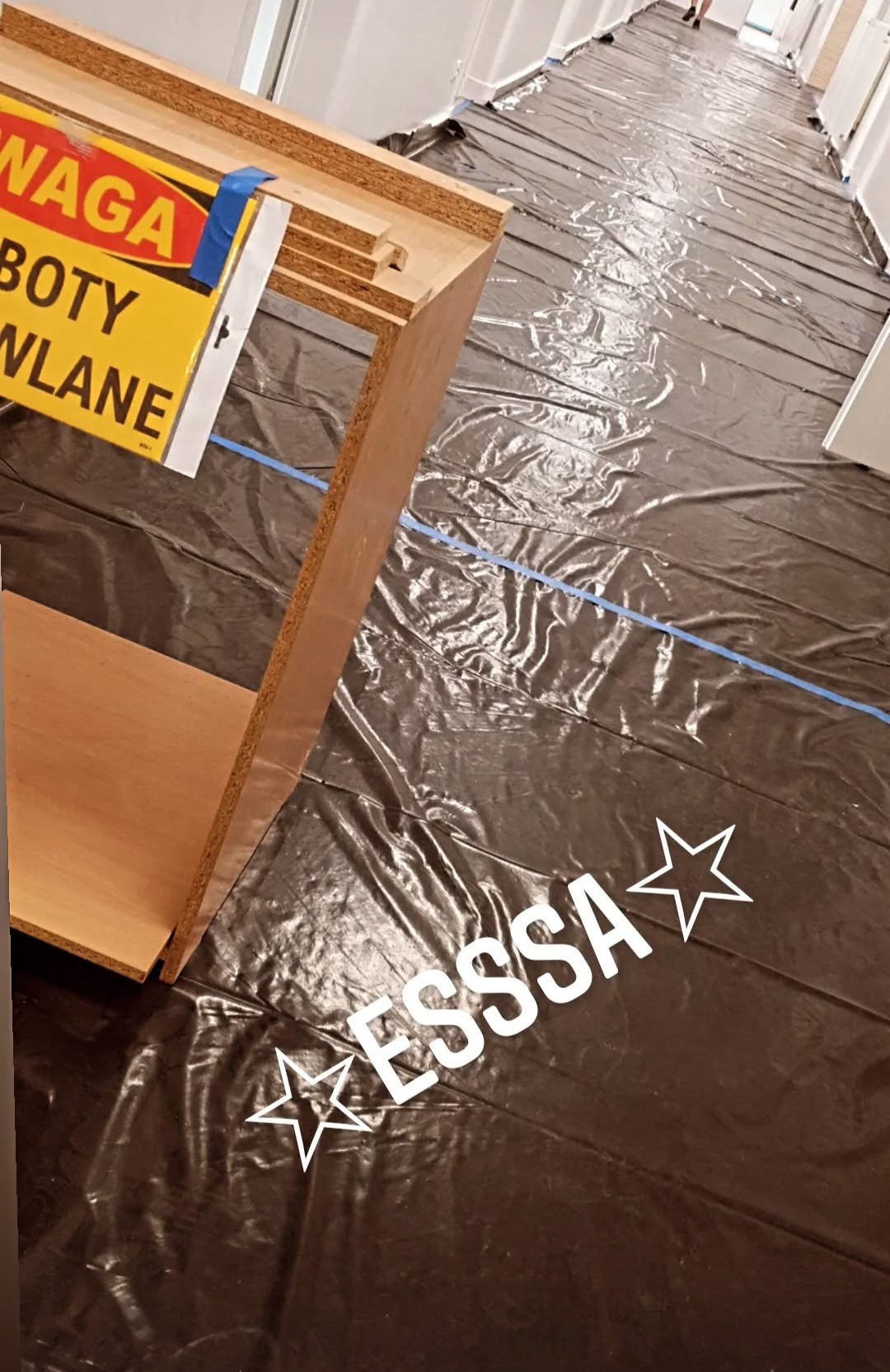 Przygotowanie do prac wykończeniowych: zabezpieczona folią podłoga w korytarzu, fragment mebla z żółtą tabliczką ostrzegawczą. Ujęcie z dołu, perspektywa zniekształcona.