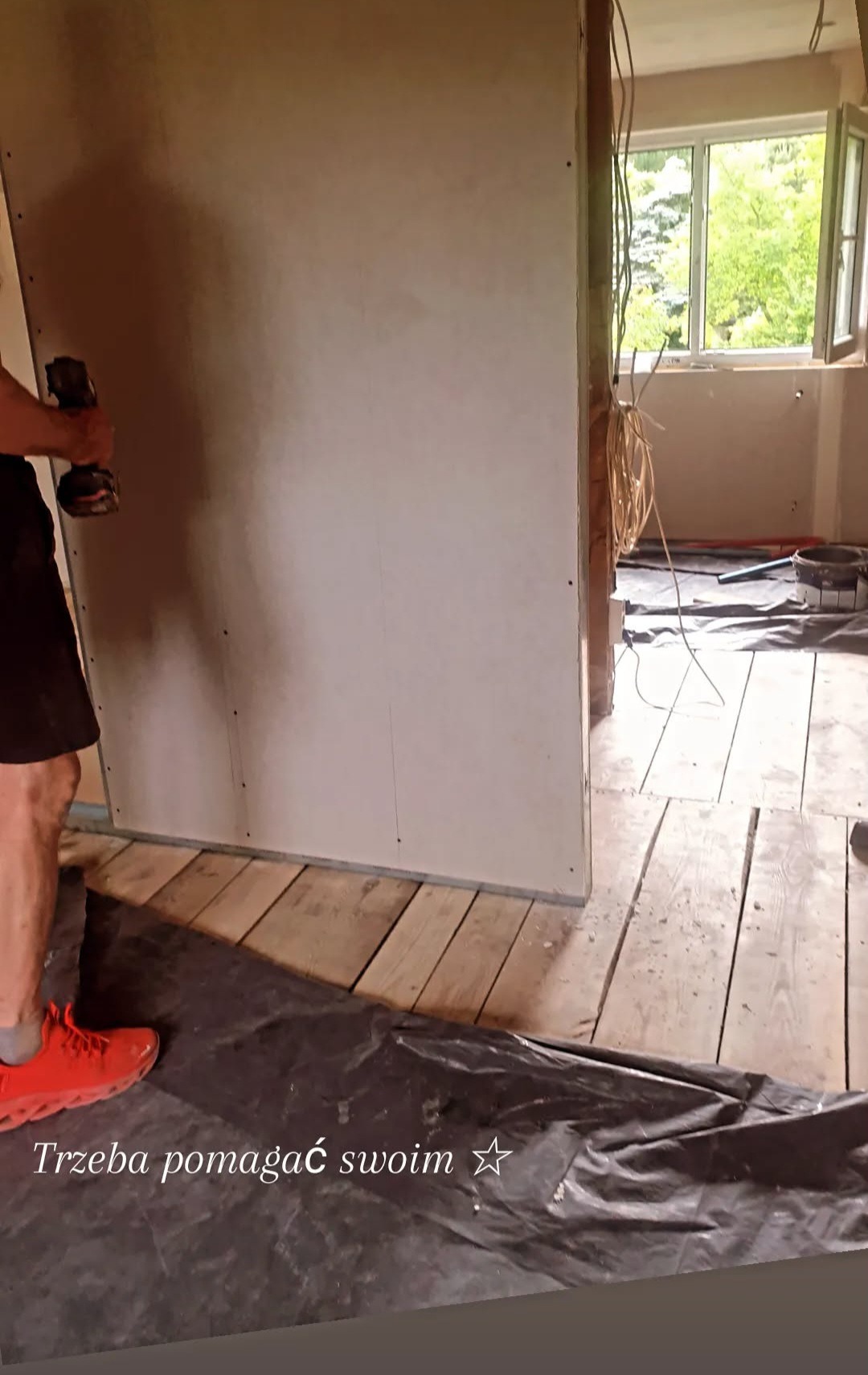 Montaż płyty gipsowej na drewnianej podłodze. Widoczne kable, okno i narzędzia. W tle zieleń. Prace wykończeniowe w trakcie realizacji.