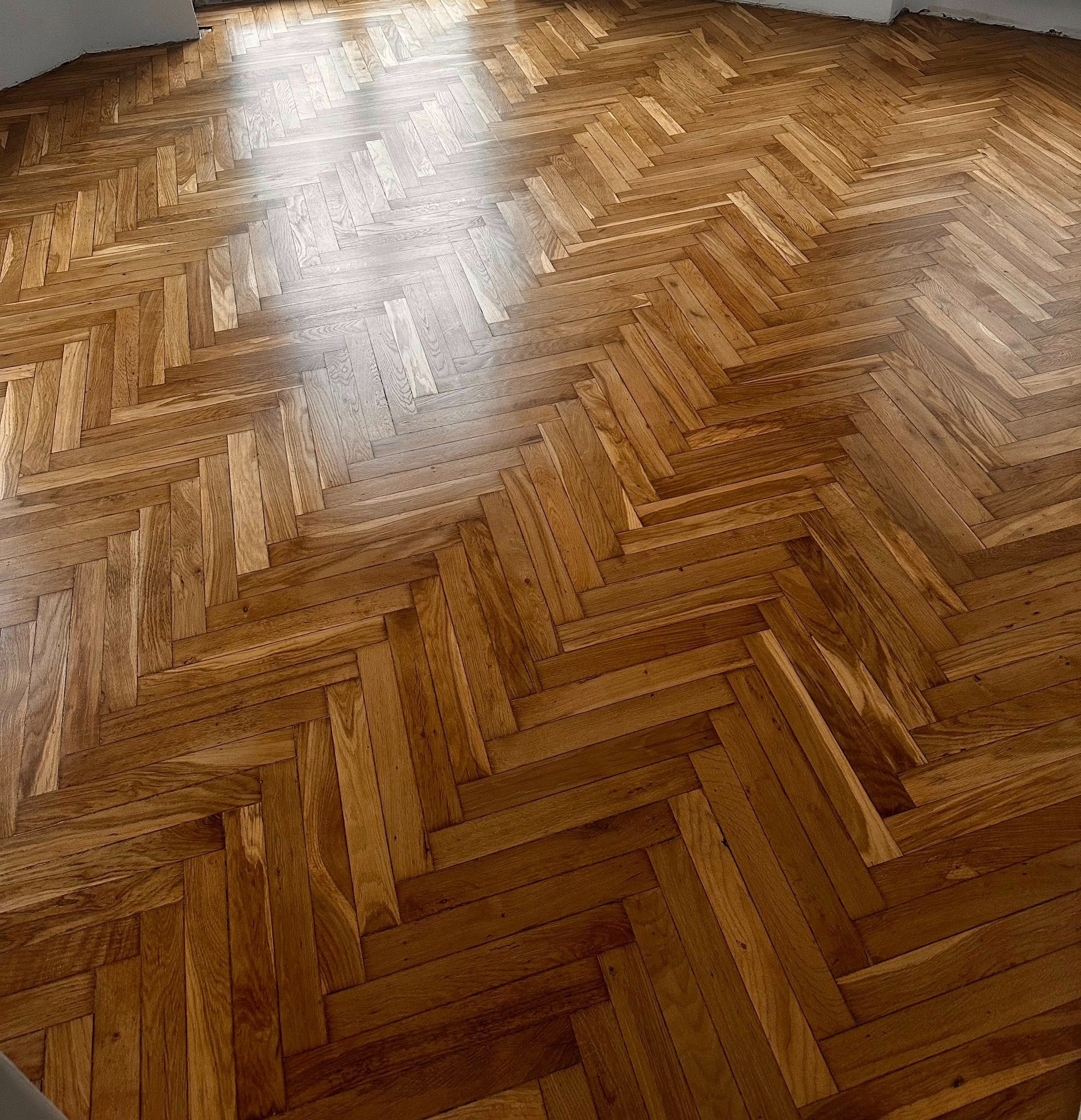 Parkiet ułożony w jodełkę po renowacji, z widocznym połyskiem i naturalnym usłojeniem drewna, oświetlony naturalnym światłem wpadającym przez okno.