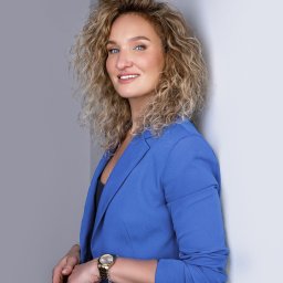 Kancelaria Adwokacka - Adwokat Marta Krajewska - Uśmiechnięta blondynka w niebieskim żakiecie, oparta o ścianę. Złoty zegarek i delikatna biżuteria dodają profesjonalizmu. Portret biznesowy.