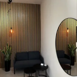 Nowoczesna poczekalnia z sofą, stolikami i ścianą z drewnianych lameli. Okrągłe lustro odbija wnętrze. Minimalistyczny design z roślinami w czarnych donicach.