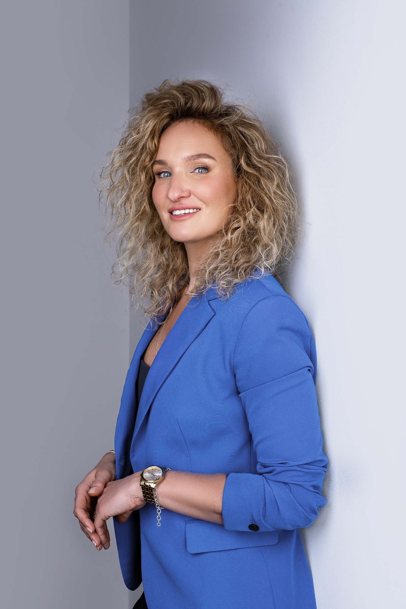 Uśmiechnięta blondynka w niebieskim żakiecie, oparta o ścianę. Złoty zegarek i delikatna biżuteria dodają profesjonalizmu. Portret biznesowy.