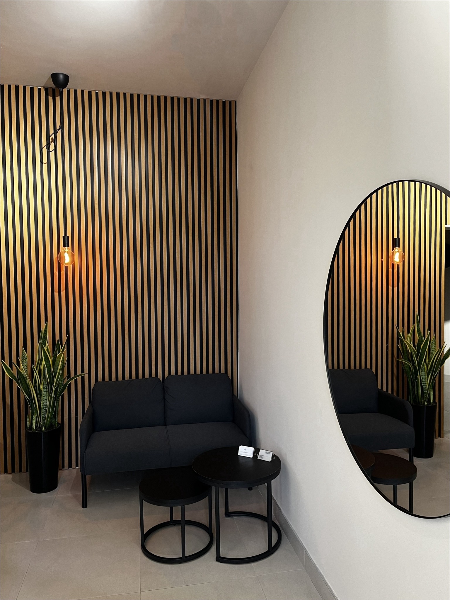 Nowoczesna poczekalnia z sofą, stolikami i ścianą z drewnianych lameli. Okrągłe lustro odbija wnętrze. Minimalistyczny design z roślinami w czarnych donicach.