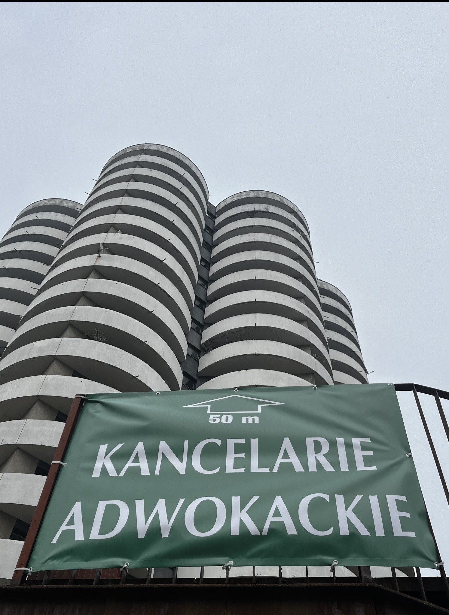 Zielony baner z napisem 'Kancelarie Adwokackie' na tle charakterystycznego budynku z cylindrycznymi balkonami w pochmurny dzień. Ujęcie z dołu.