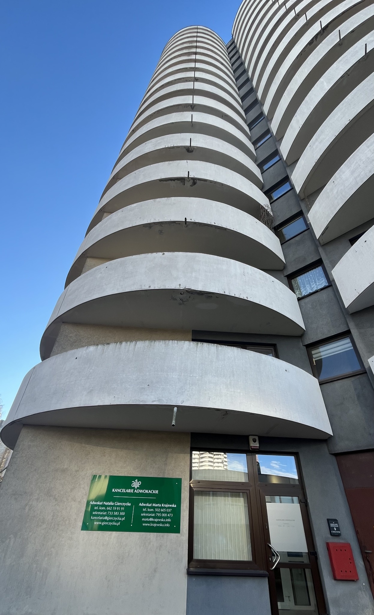 Wejście do kancelarii adwokackiej w charakterystycznym, okrągłym budynku z balkonami. Tabliczka informacyjna z nazwiskami adwokatów i danymi kontaktowymi.