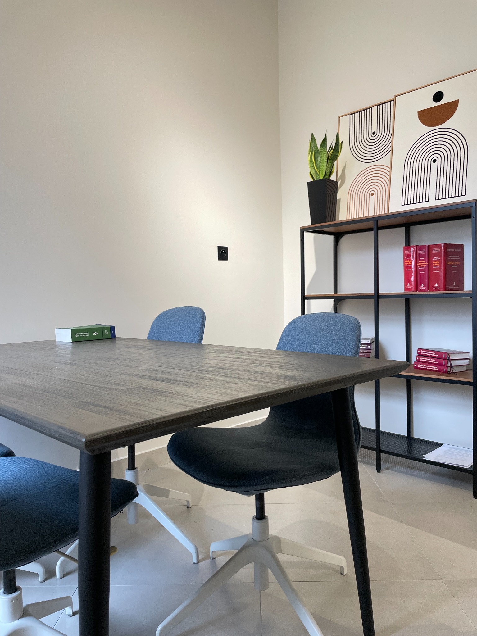 Nowoczesne biuro z drewnianym stołem, krzesłami na kółkach i regałem z książkami prawniczymi. Na stole leży zielony kodeks. Minimalistyczne obrazy na ścianie.