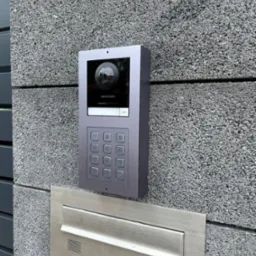 Nowoczesny wideodomofon Hikvision z klawiaturą numeryczną, zamontowany na szarej, betonowej ścianie obok metalowej skrzynki na listy.