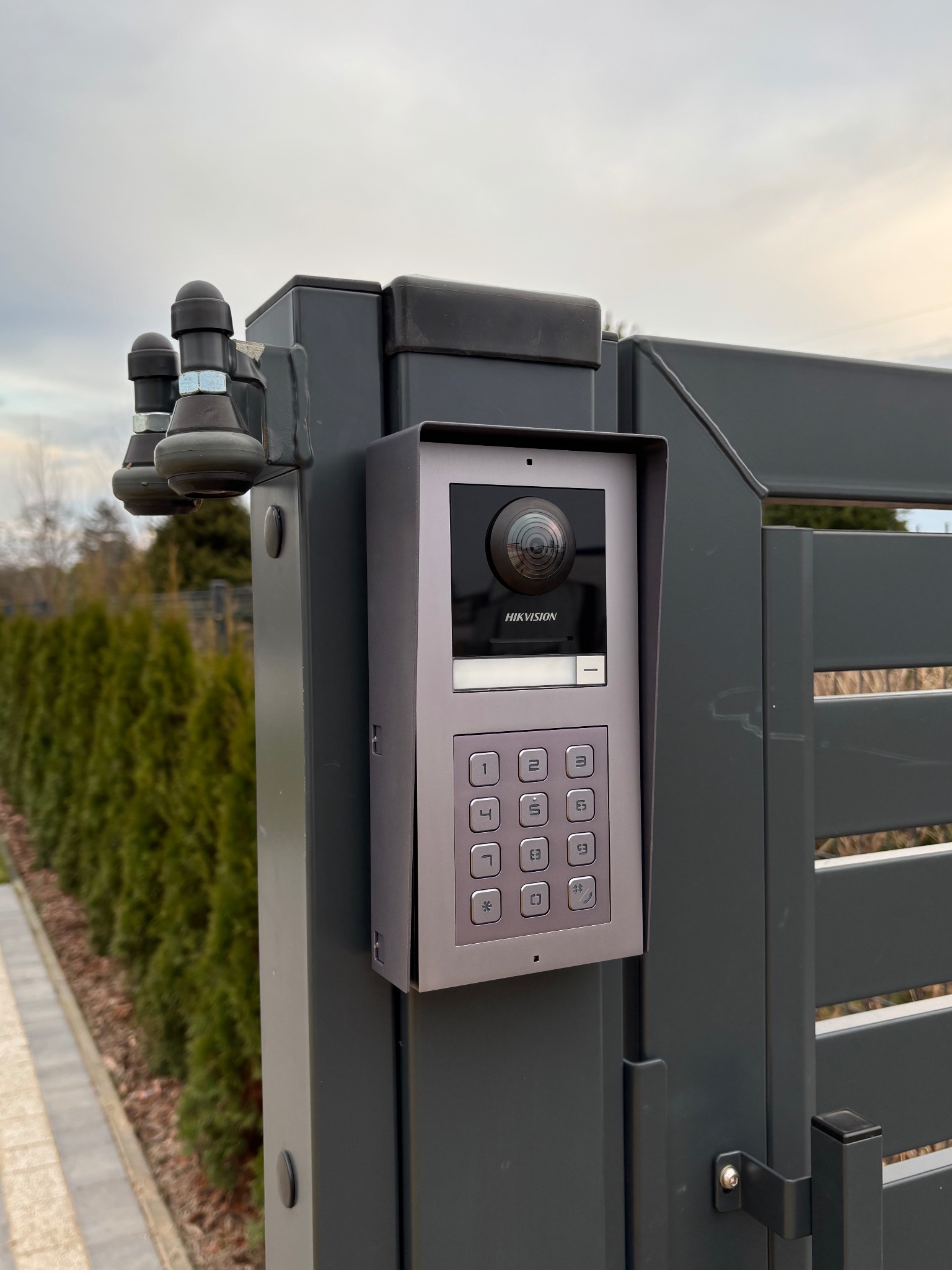 Szary wideodomofon Hikvision zamontowany na metalowej bramie posesji, z widocznymi przyciskami numerycznymi i kamerą. W tle zieleń i fragment chodnika.