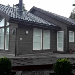 Drewniany dom z ciemną elewacją i dużymi oknami, otoczony lasem. Widoczny taras z drewnianych desek i elementy oświetlenia zewnętrznego. Dach pokryty ciemną dachówką.