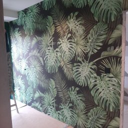 A.F. KOMFORT ADAM FIGURSKI - Ściana z fototapetą w tropikalne liście monstery i palmy, widoczny fragment sufitu z lampą, podłoga przygotowana do dalszych prac. Fragment drabiny w rogu.