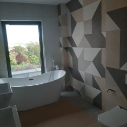 A.F. KOMFORT ADAM FIGURSKI - Nowoczesna łazienka z wolnostojącą wanną przy oknie z widokiem na zieleń. Ściana z geometrycznym wzorem z płytek w odcieniach szarości i beżu. Minimalistyczna toaleta.