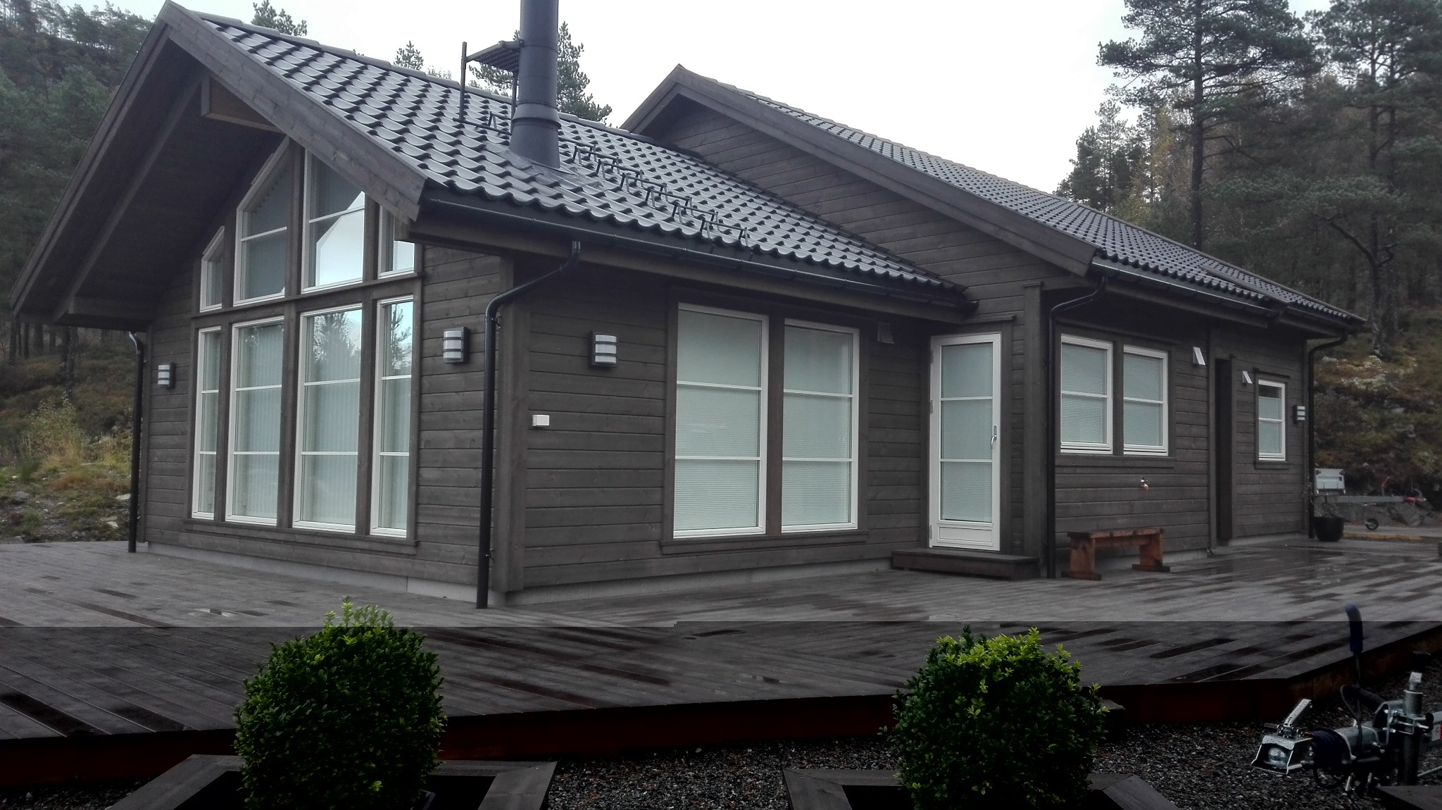 Drewniany dom z ciemną elewacją i dużymi oknami, otoczony lasem. Widoczny taras z drewnianych desek i elementy oświetlenia zewnętrznego. Dach pokryty ciemną dachówką.