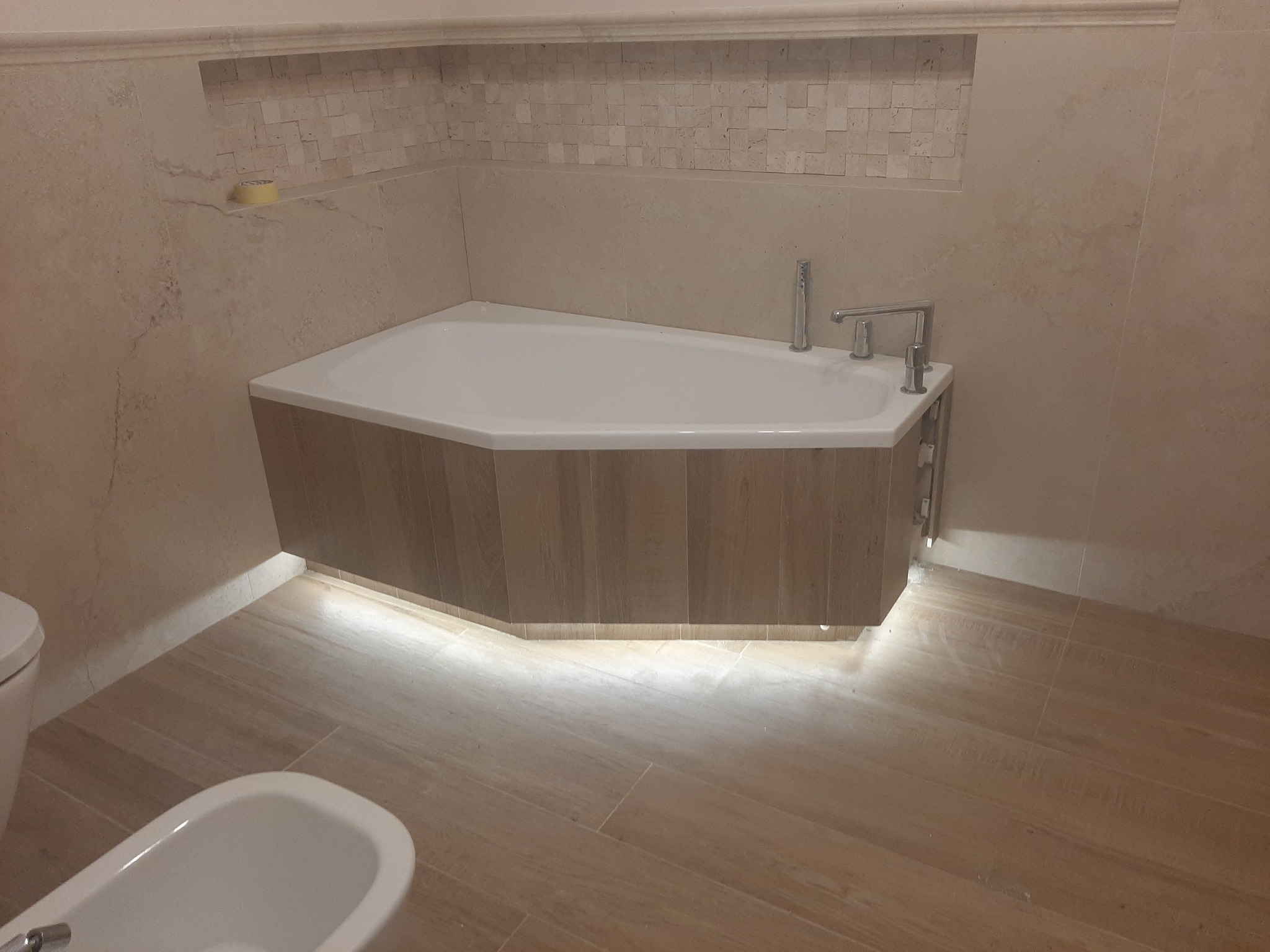 Wykończona łazienka z narożną wanną obudowaną drewnem, podświetleniem LED i beżowymi płytkami imitującymi drewno na podłodze i ścianach. Widoczny bidet i mozaika w niszy.