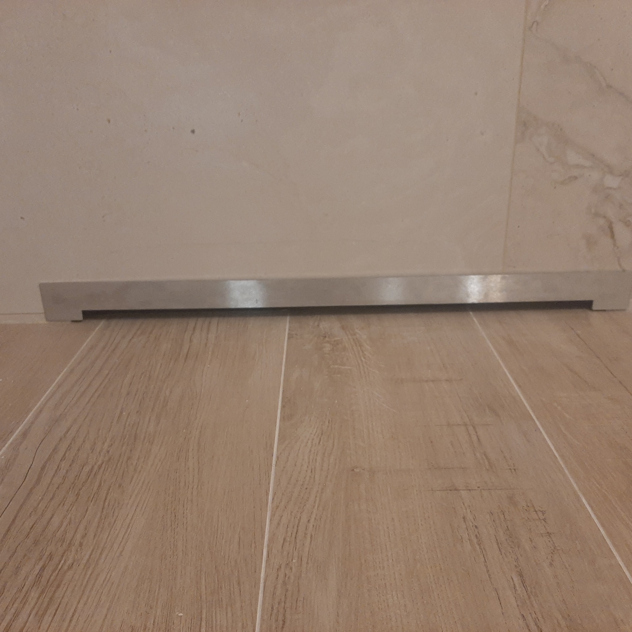 Ułożona podłoga z paneli imitujących drewno, przy ścianie zamontowana srebrna listwa przypodłogowa. Jasne płytki na ścianie w tle.