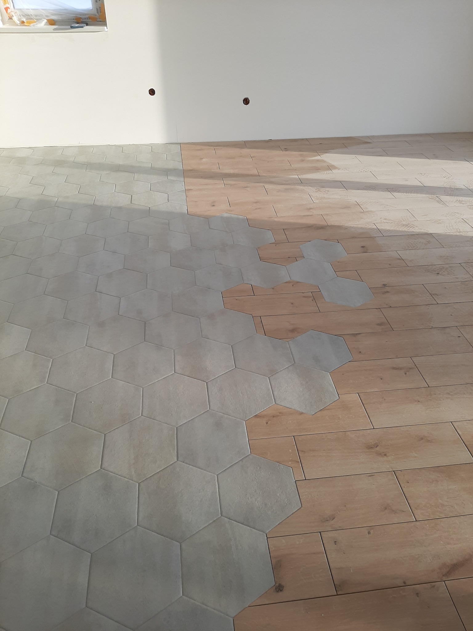 Układanie heksagonalnych płytek podłogowych w jasnoszarym odcieniu, częściowo na drewnopodobnej powierzchni. Widoczne postępy prac w jasnym pomieszczeniu.