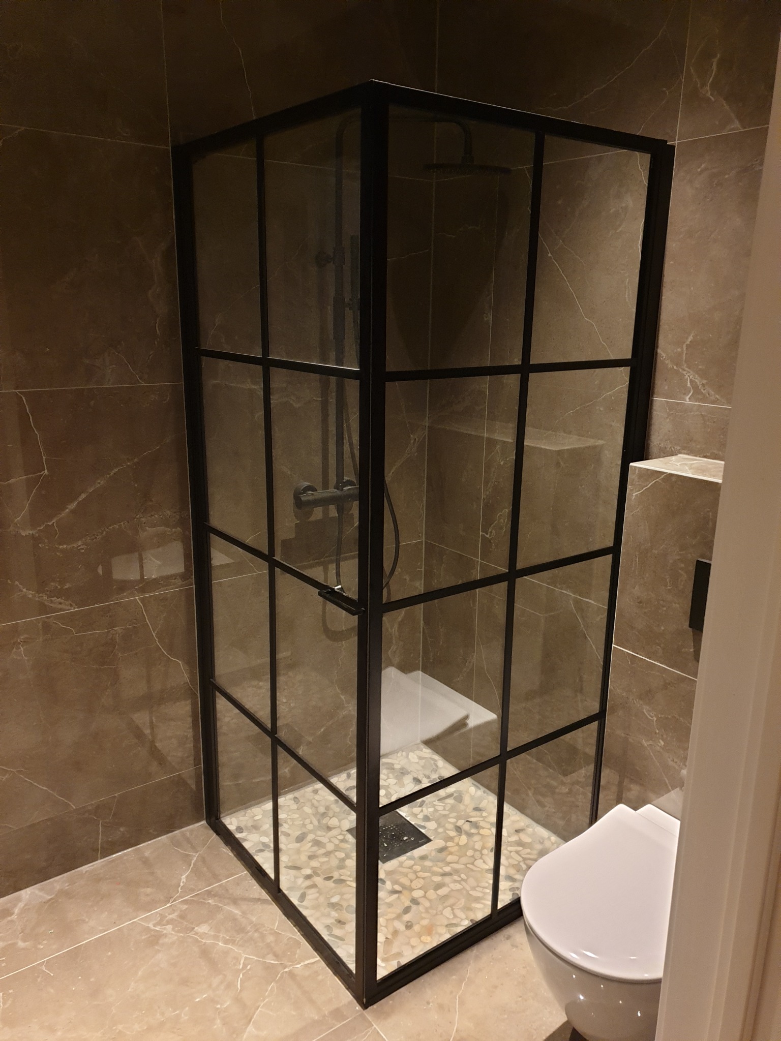 Nowoczesna kabina prysznicowa z czarnymi profilami i kamienną posadzką w łazience wykończonej płytkami imitującymi marmur. Widoczna toaleta i odpływ liniowy.
