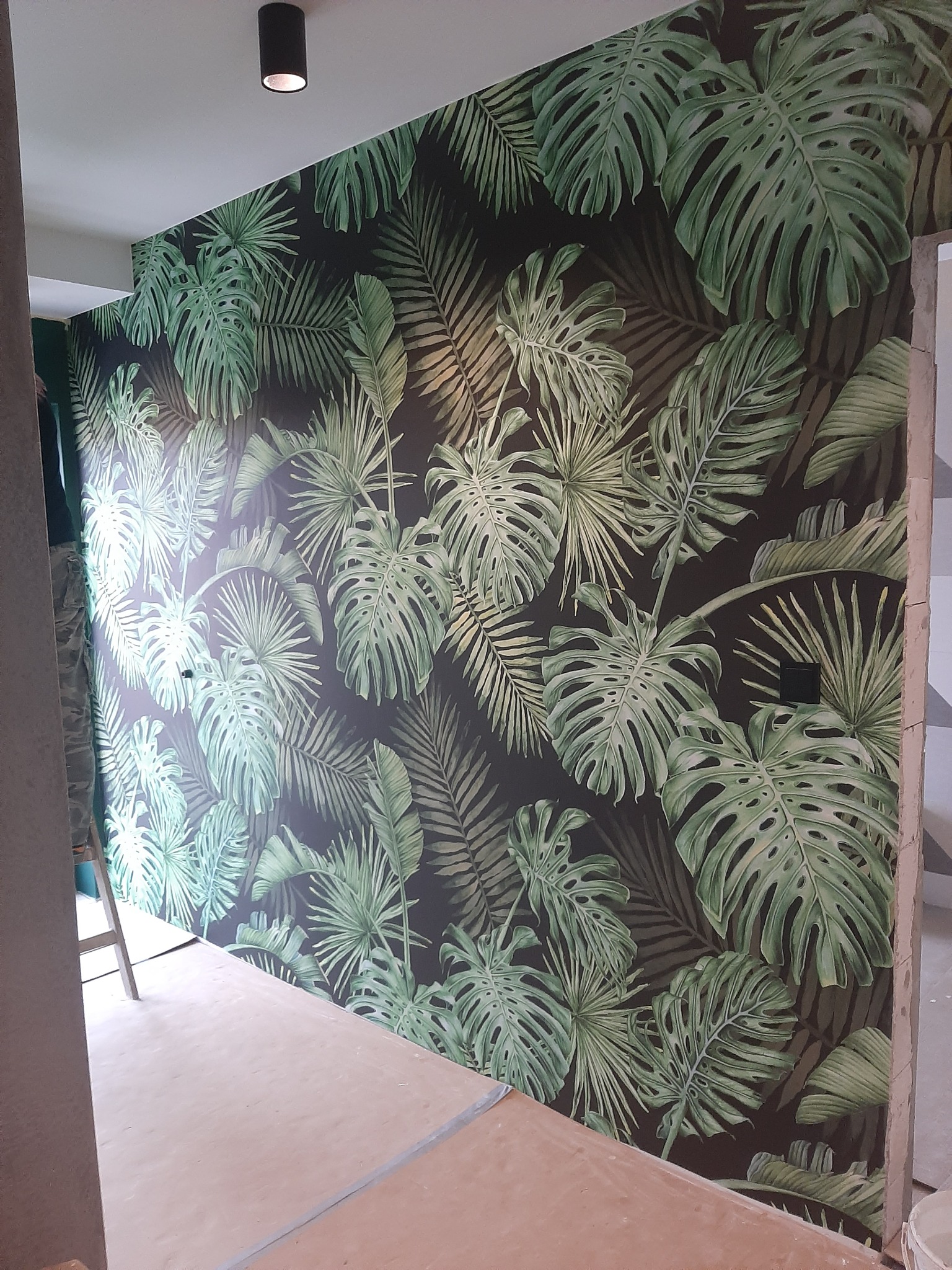 Ściana z fototapetą w tropikalne liście monstery i palmy, widoczny fragment sufitu z lampą, podłoga przygotowana do dalszych prac. Fragment drabiny w rogu.