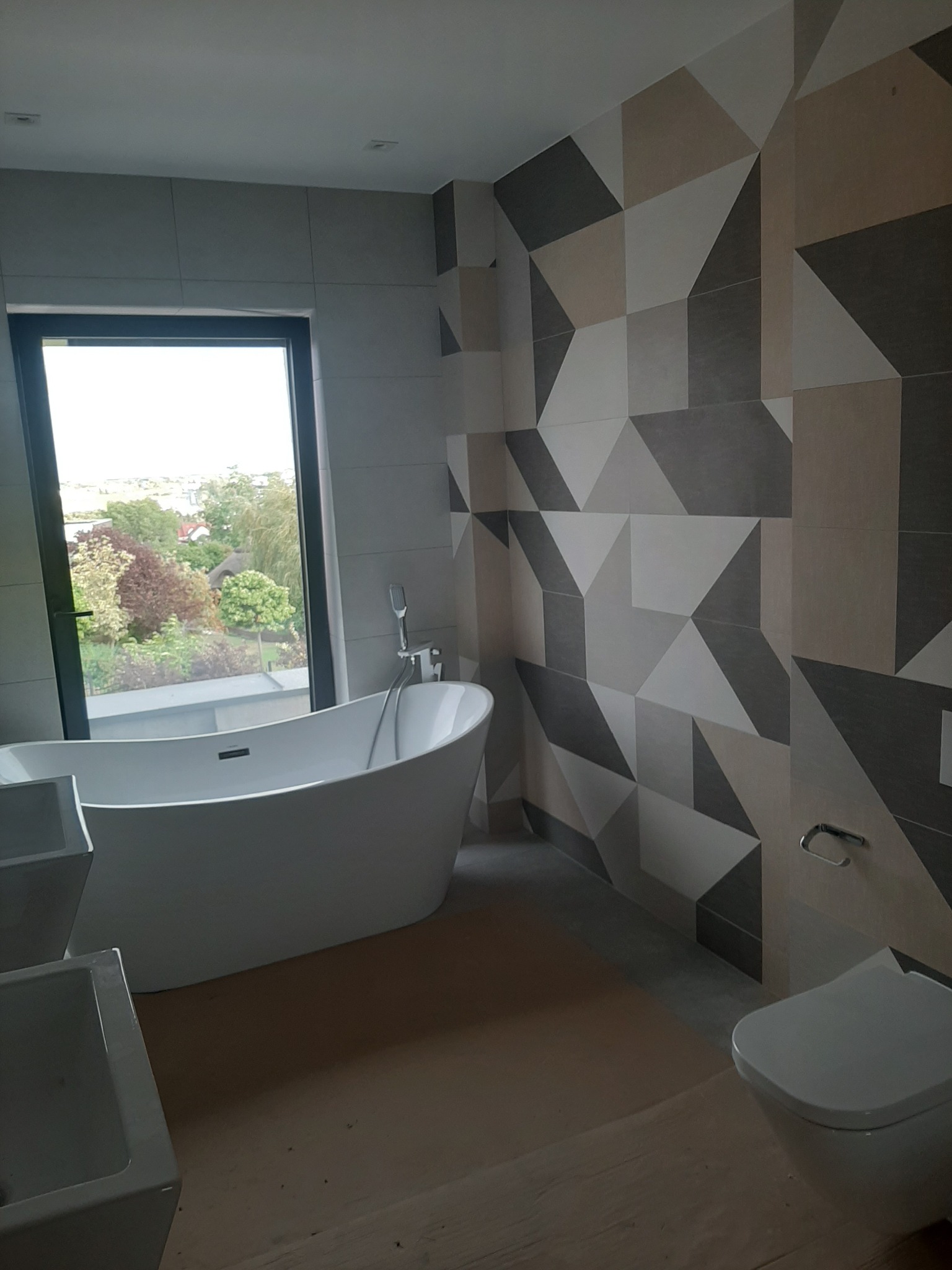 Nowoczesna łazienka z wolnostojącą wanną przy oknie z widokiem na zieleń. Ściana z geometrycznym wzorem z płytek w odcieniach szarości i beżu. Minimalistyczna toaleta.