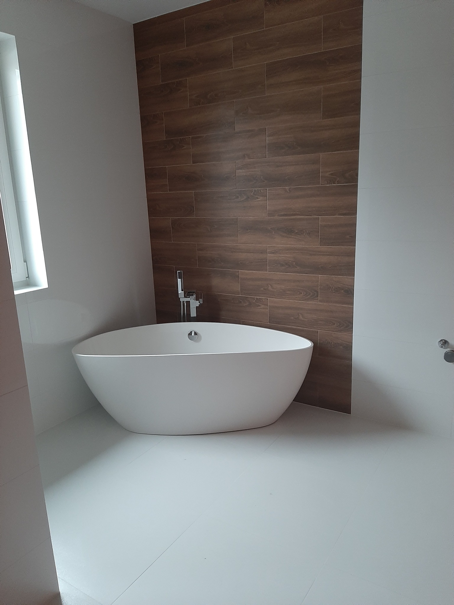 Nowoczesna łazienka z wolnostojącą wanną na białej podłodze. Ściana wyłożona płytkami imitującymi drewno. Minimalistyczna armatura i jasne wnętrze.