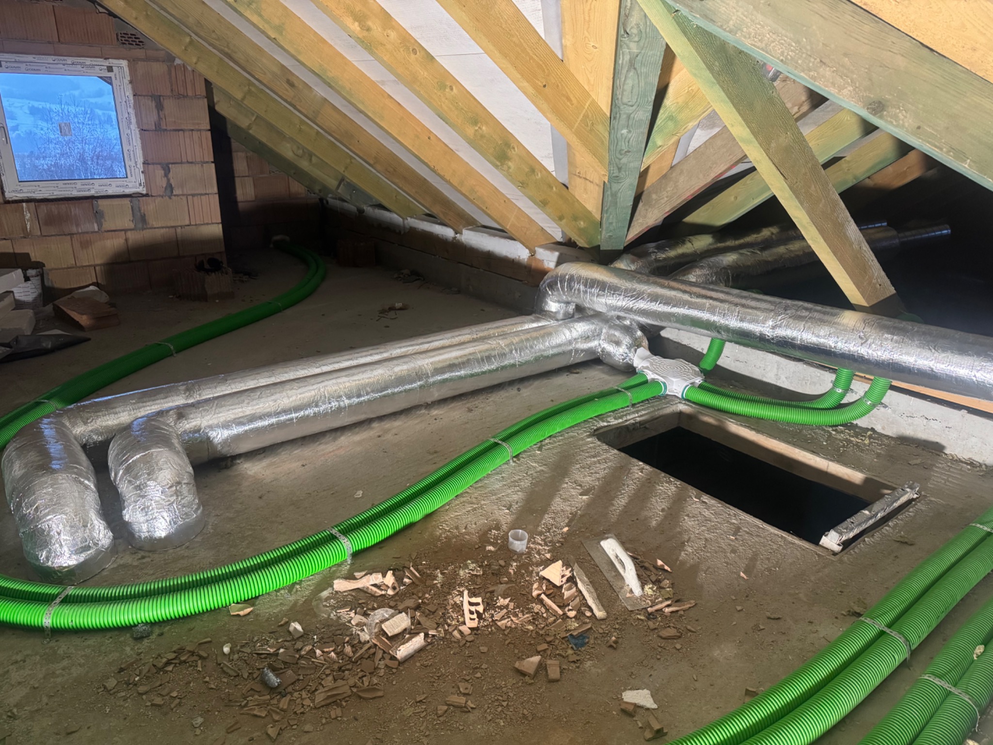 Instalacja rekuperacji na poddaszu w Limanowej. Widoczne srebrne rury izolowane i zielone przewody wentylacyjne. W tle okno i drewniana konstrukcja dachu.