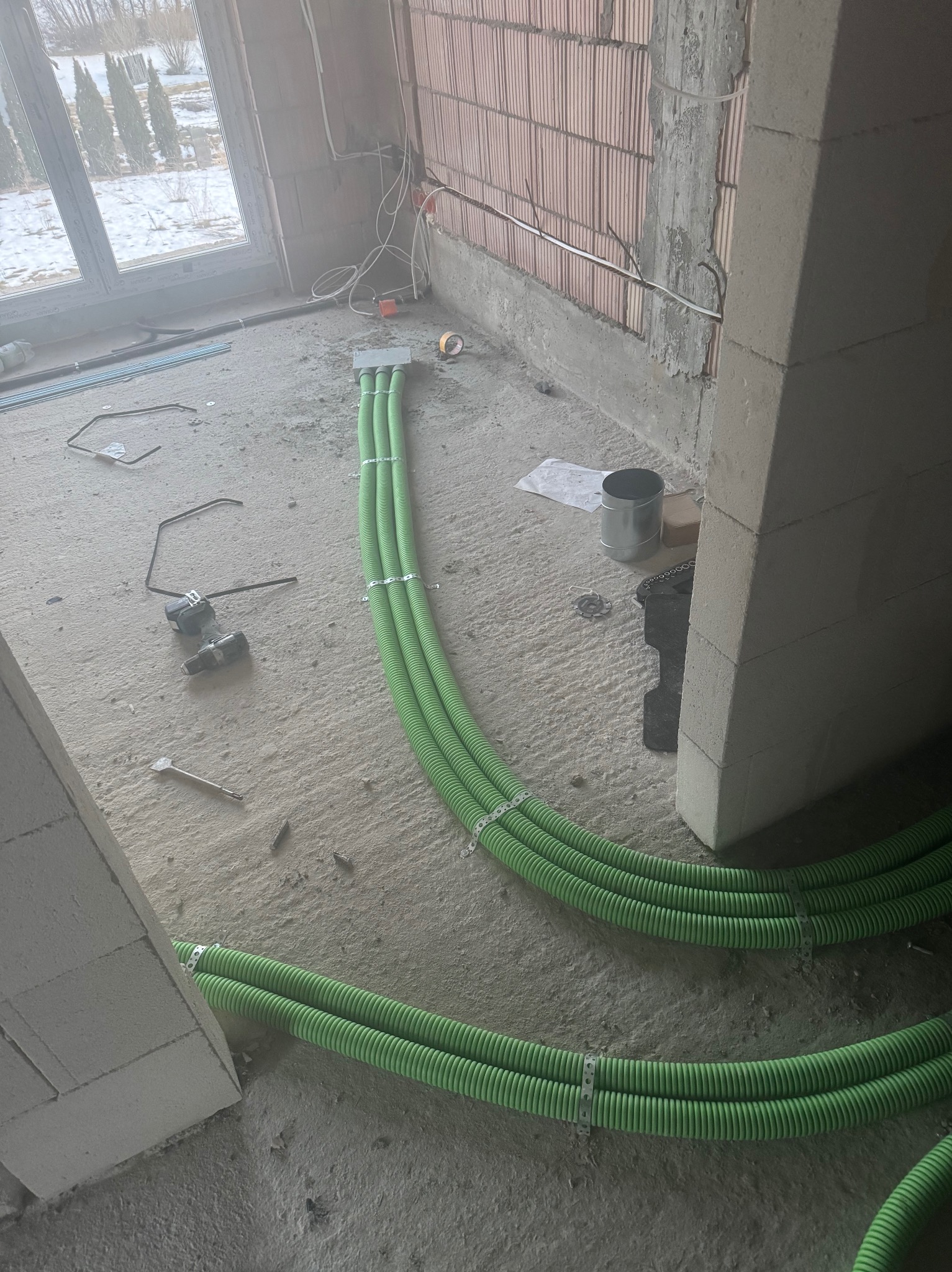 Zielone rury wentylacyjne, ułożone równolegle na surowym podłożu w trakcie montażu w budynku. Widoczne narzędzia i elementy instalacji elektrycznej.