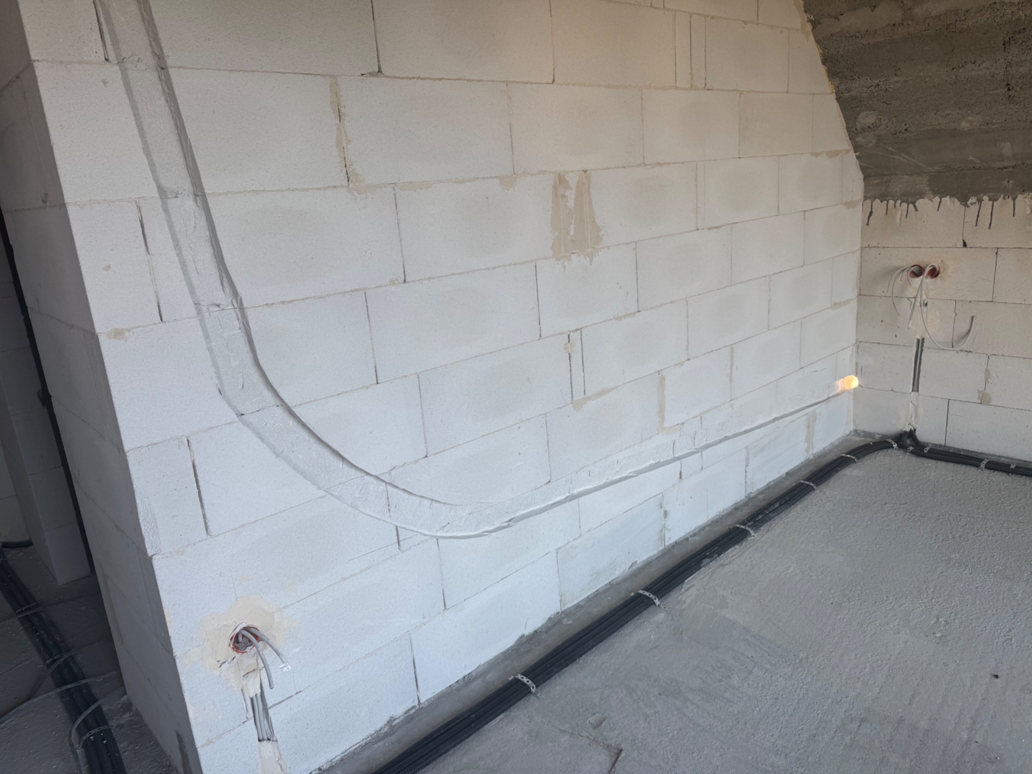 Instalacja przewodów wentylacyjnych w bruzdach ściany z betonu komórkowego. Widoczne kable ułożone wzdłuż podłogi, przymocowane metalowymi uchwytami.