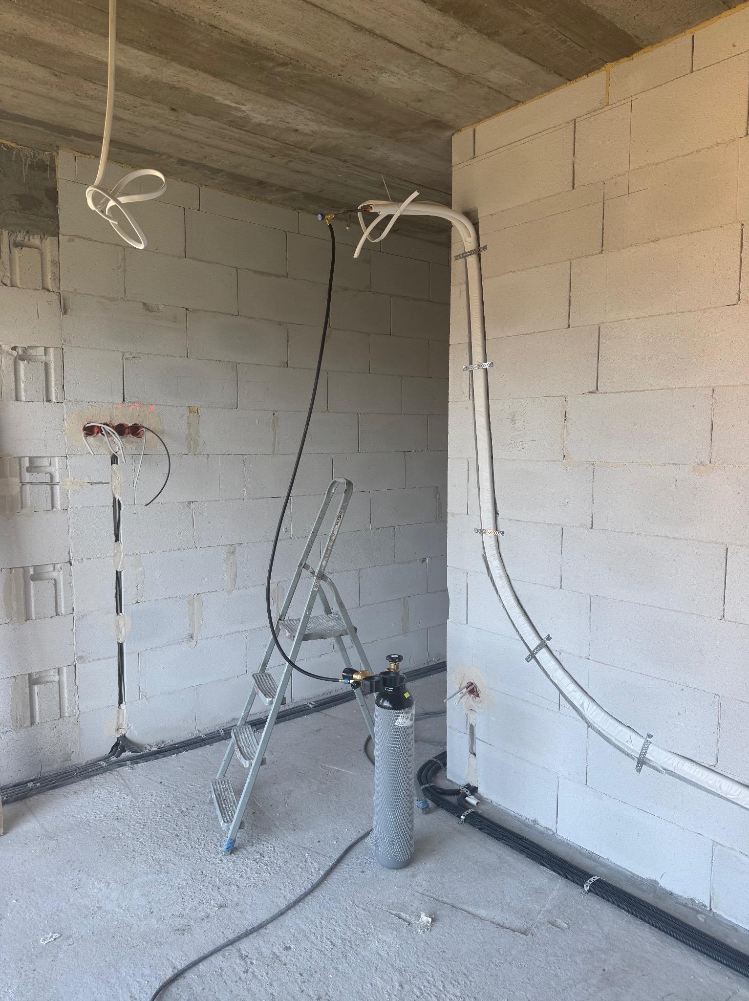 Surowy stan: montaż instalacji wentylacyjnej w budynku z białego bloczka. Widoczne rury, przewody, drabina i butla z gazem technicznym. Etap prac instalacyjnych.