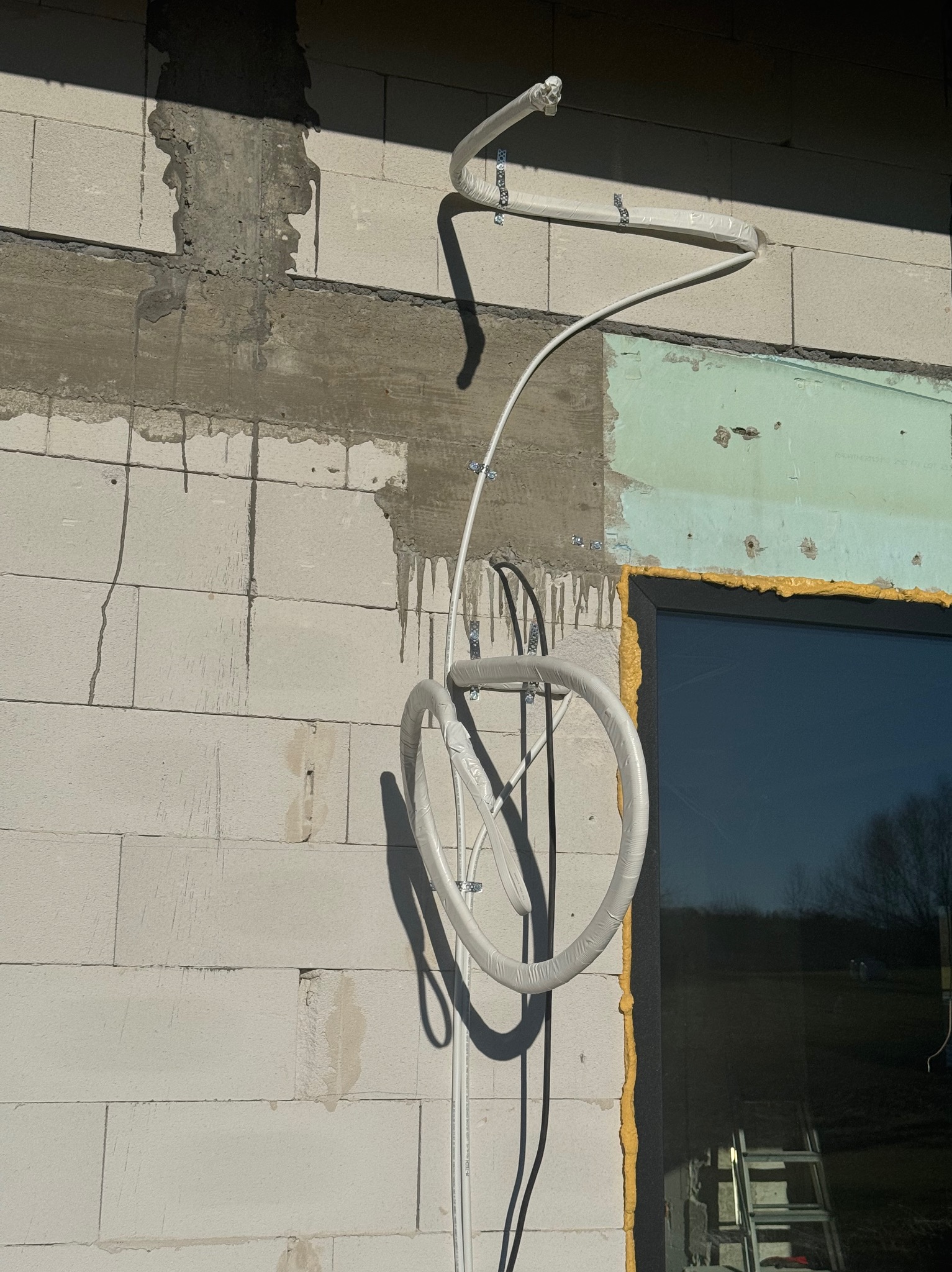 Białe rury wentylacyjne zamontowane na surowej ścianie z betonu komórkowego, widoczne mocowania i fragment okna z żółtą pianką montażową.