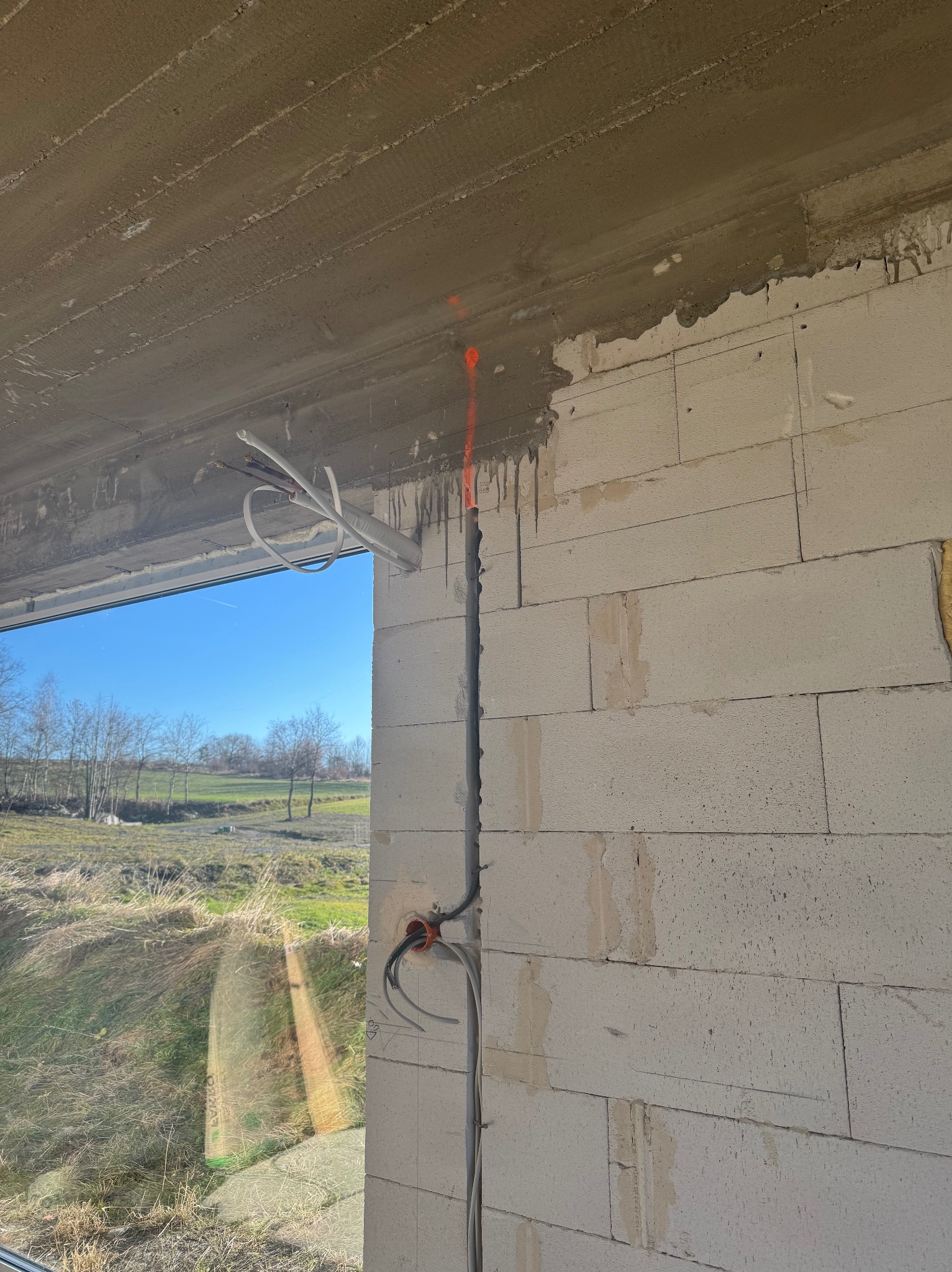 Montaż instalacji: przewody i rury prowadzone w ścianie z betonu komórkowego, widok przez okno na zewnątrz. Etap prac instalacyjnych w surowym stanie.
