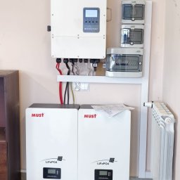 WARMEKO SPÓŁKA Z OGRANICZONĄ ODPOWIEDZIALNOŚCIĄ - Biała instalacja fotowoltaiczna MUST z akumulatorami LiFePO4 na ścianie, widoczne kable i zabezpieczenia. Element grzejnika z boku.