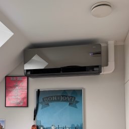 WARMEKO SPÓŁKA Z OGRANICZONĄ ODPOWIEDZIALNOŚCIĄ - Czarny klimatyzator ścienny Versu Rotenso z widocznym wyświetlaczem temperatury 16.0, zamontowany w pokoju z plakatami Bon Jovi. Białe rury instalacyjne.