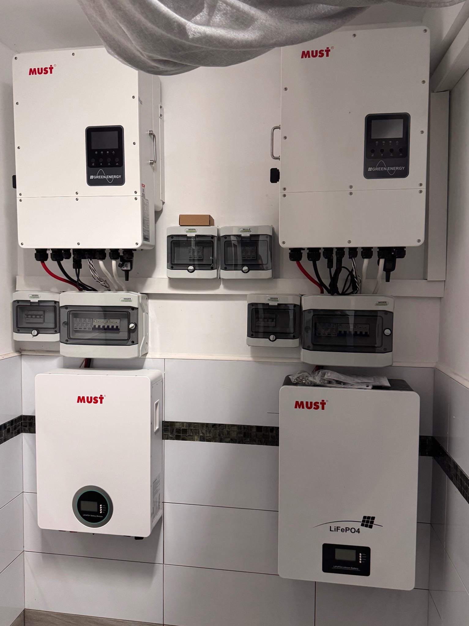 Kompletna instalacja fotowoltaiczna w pomieszczeniu: białe inwertery MUST, rozdzielnice elektryczne NOARK, kable. Układ na kafelkowanej ścianie.