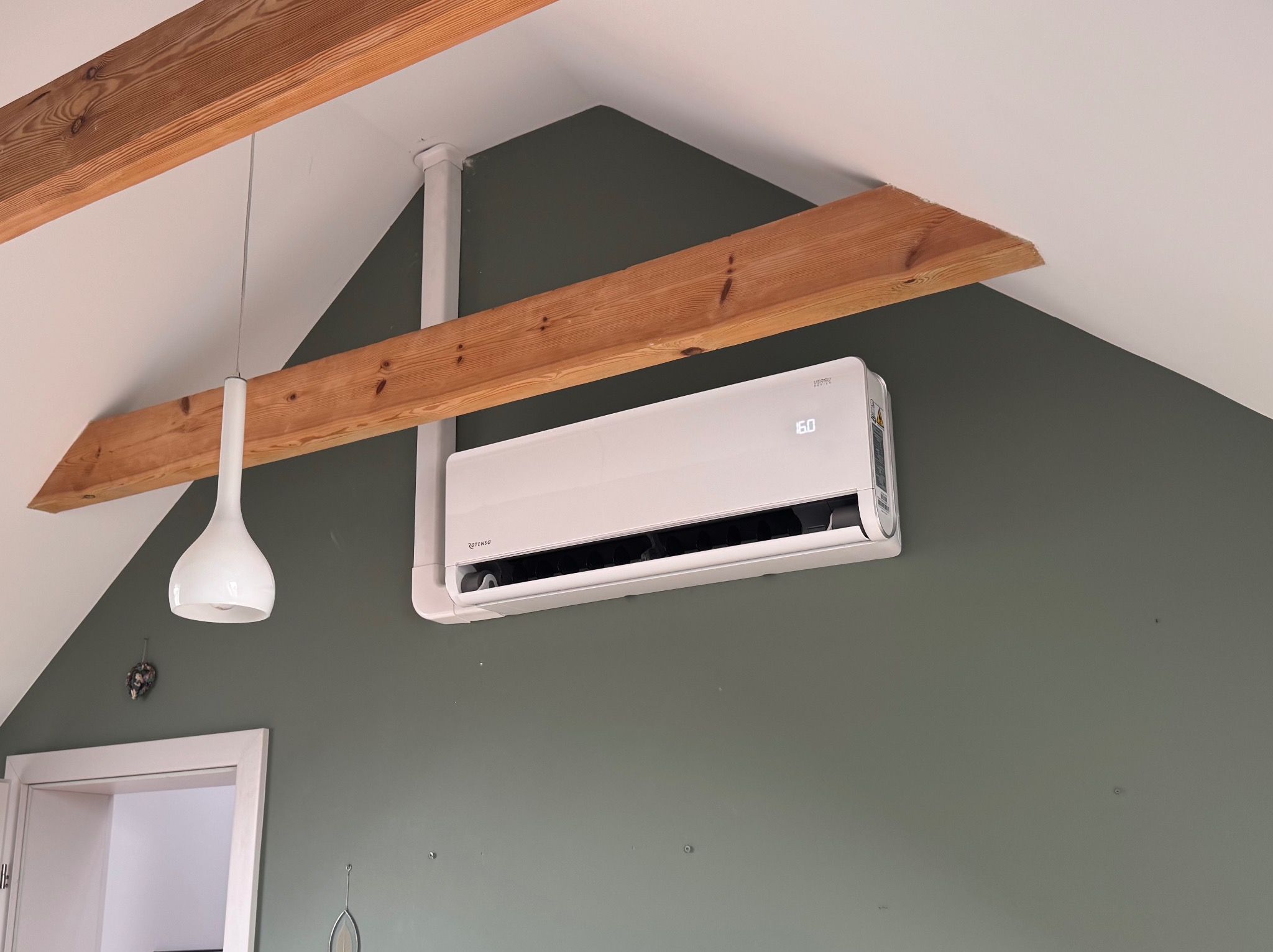Biały klimatyzator ścienny marki Rotenso zamontowany w pomieszczeniu z drewnianymi belkami stropowymi i lampą wiszącą. Widok na szarozieloną ścianę.