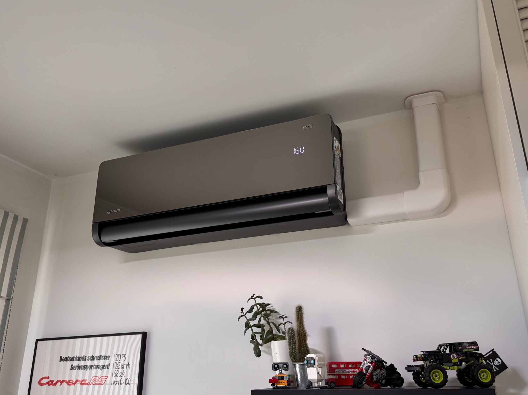 Czarny klimatyzator Rotenso Versu z wyświetlaczem 16.0, zamontowany na ścianie w pokoju. Białe rury instalacyjne. Na półce dekoracje i plakat Carrera RS.