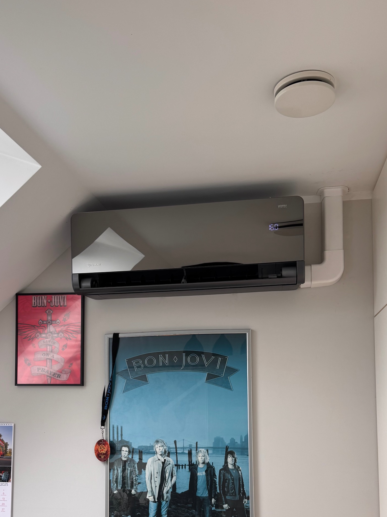 Czarny klimatyzator ścienny Versu Rotenso z widocznym wyświetlaczem temperatury 16.0, zamontowany w pokoju z plakatami Bon Jovi. Białe rury instalacyjne.