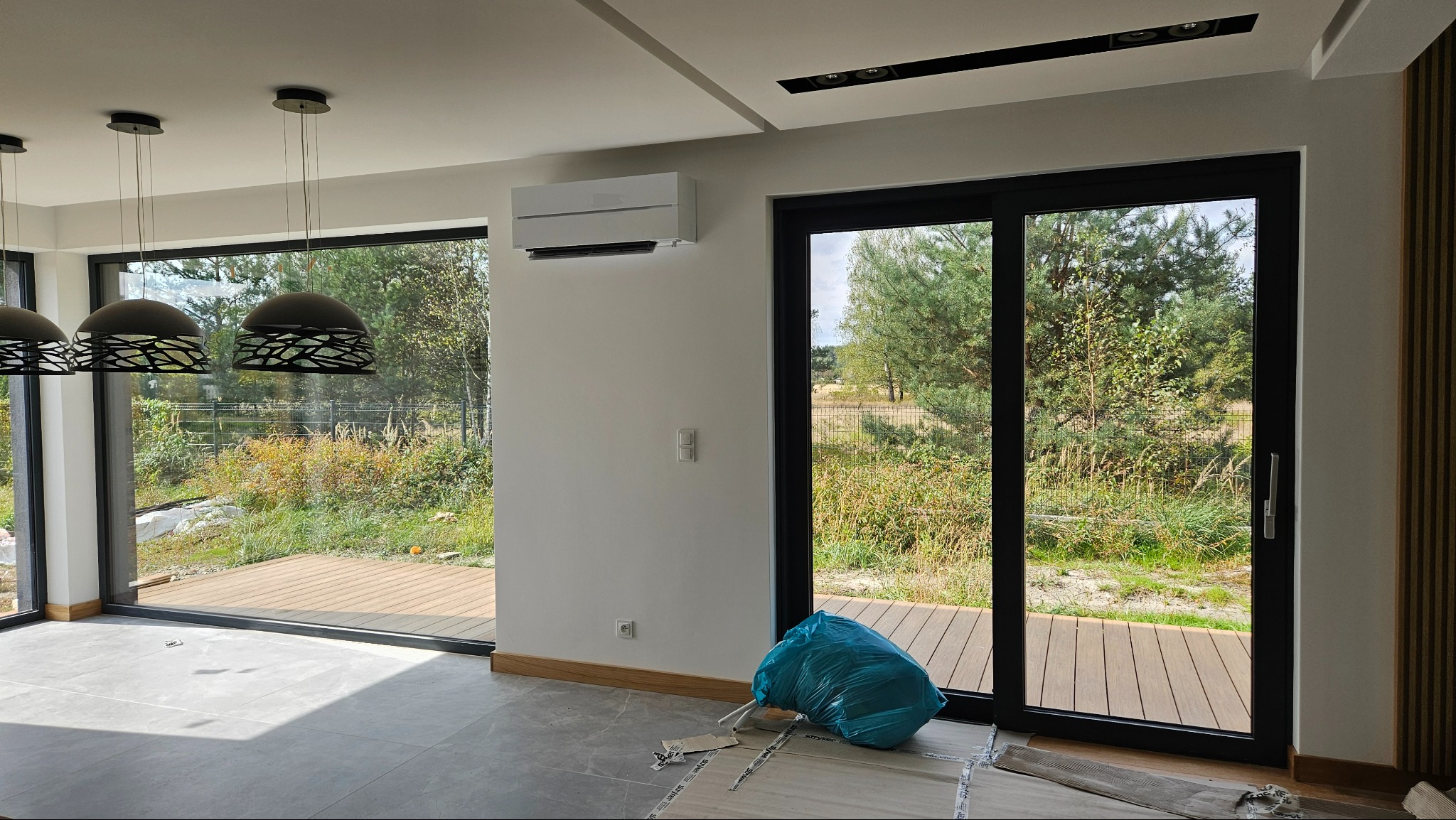 Nowoczesne wnętrze z klimatyzatorem Mitsubishi na ścianie, widok na zewnątrz przez duże okna. Na podłodze folia zabezpieczająca, worek z odpadami.
