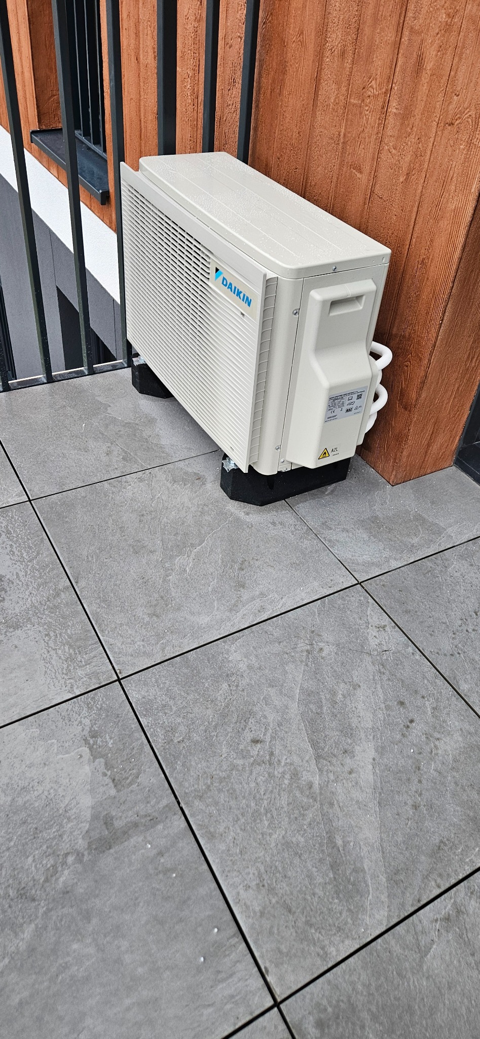 Biała jednostka zewnętrzna klimatyzacji Daikin na balkonie, zamontowana na czarnych podkładkach antywibracyjnych. Widoczne rury i fragment drewnianej ściany.