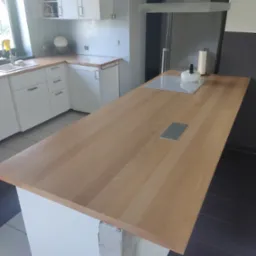 Jasny, drewniany blat kuchenny z gniazdkami, zamontowany na białej wyspie kuchennej. W tle widoczne białe szafki i okap. Uwagę zwraca surowy betonowy fragment podstawy wyspy.