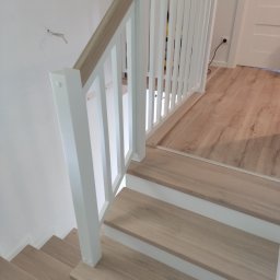 DREW-KAM - Jasne, drewniane stopnie z białą podstopnicą i białą balustradą z drewnianym poręczą. Widok z góry na dół, z fragmentem podłogi w tle.