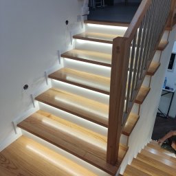 DREW-KAM - Nowoczesne schody z podświetleniem LED, drewniane stopnie o jasnym odcieniu, metalowa balustrada. Widok z dołu, oświetlenie podkreśla strukturę drewna.