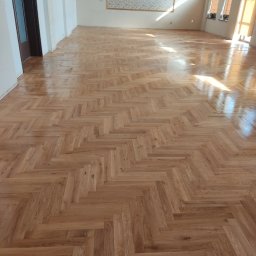 DREW-KAM - Parkiet ułożony w jodełkę w przestronnym, jasnym wnętrzu. Widoczne naturalne usłojenie drewna i połysk lakieru. W tle okna i drzwi.