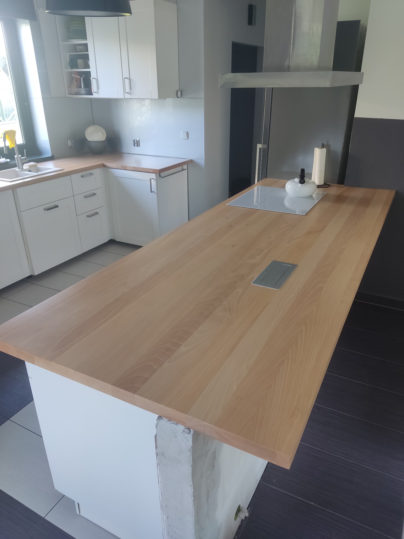 Jasny, drewniany blat kuchenny z gniazdkami, zamontowany na białej wyspie kuchennej. W tle widoczne białe szafki i okap. Uwagę zwraca surowy betonowy fragment podstawy wyspy.