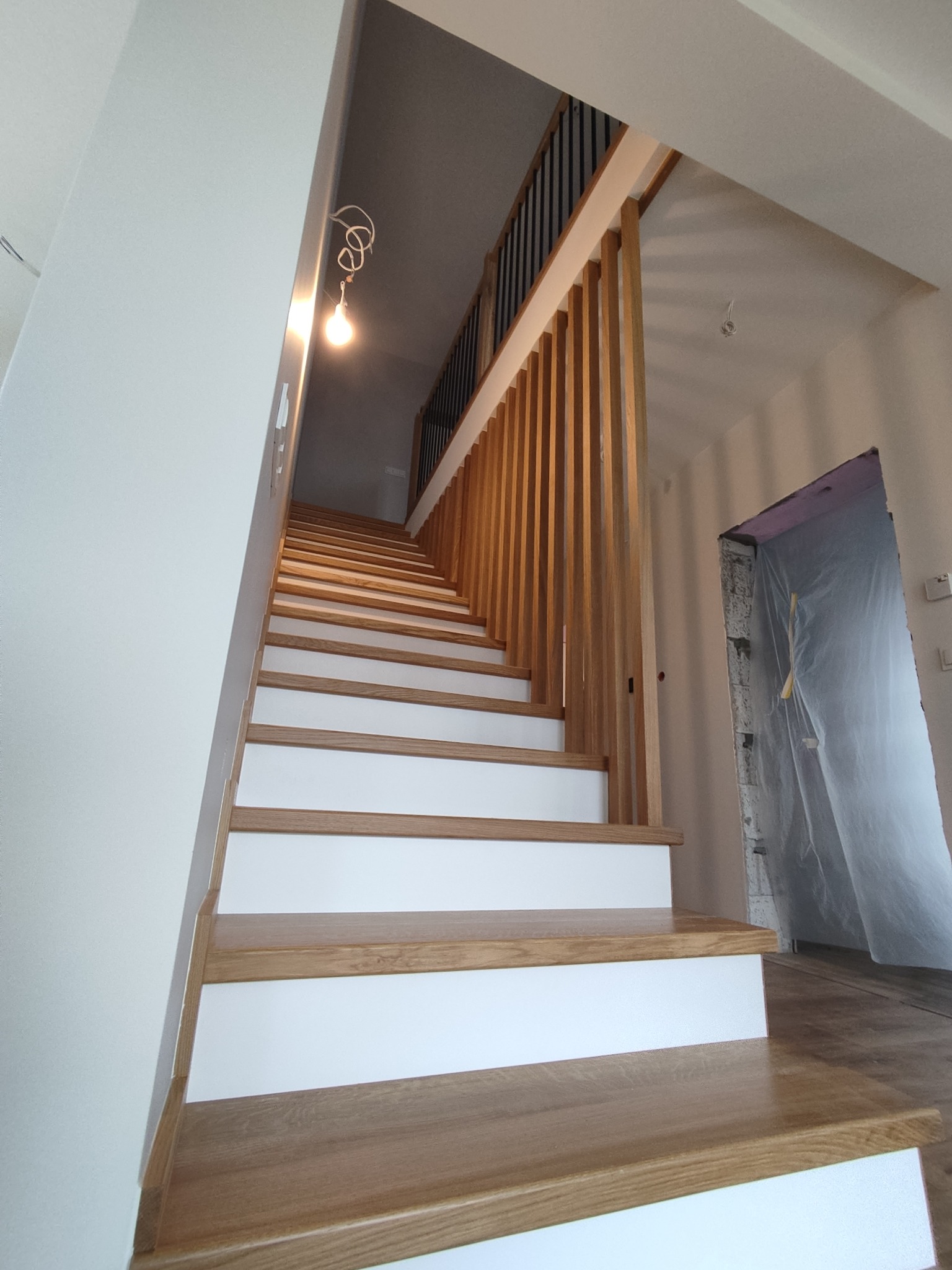 Nowoczesne schody z drewnianymi stopniami i białymi podstopnicami, z ażurową balustradą. Widok z dołu, w tle zabezpieczone folią drzwi.