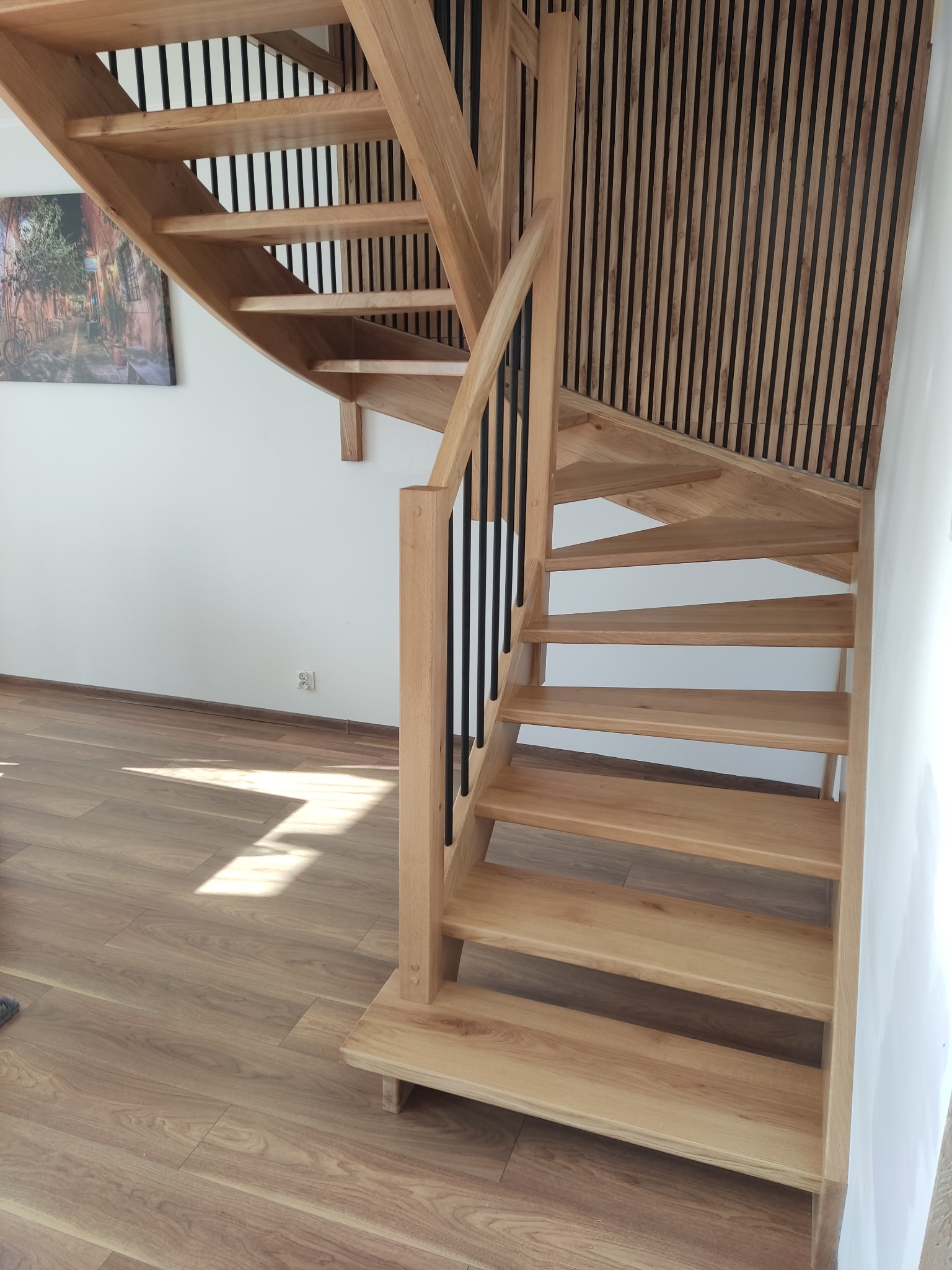 Spiralne, jasne schody z drewnianymi stopniami i czarną balustradą, z widoczną dekoracyjną ścianą z drewnianych listew i fragmentem obrazu na ścianie.