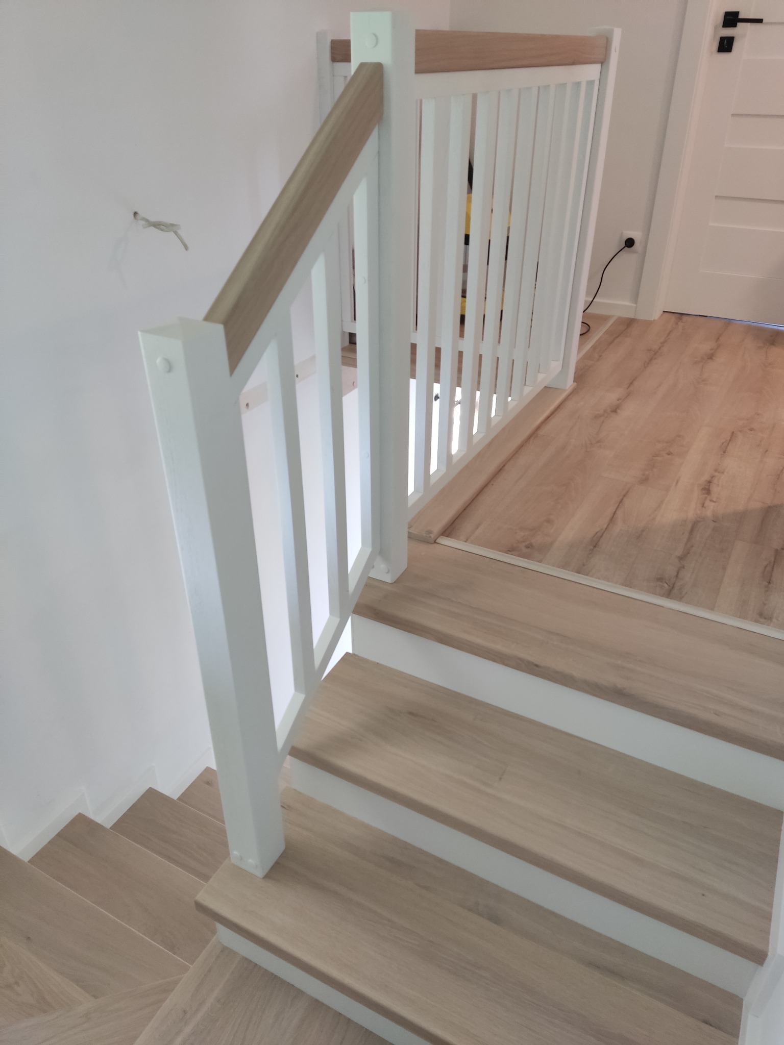 Jasne, drewniane stopnie z białą podstopnicą i białą balustradą z drewnianym poręczą. Widok z góry na dół, z fragmentem podłogi w tle.