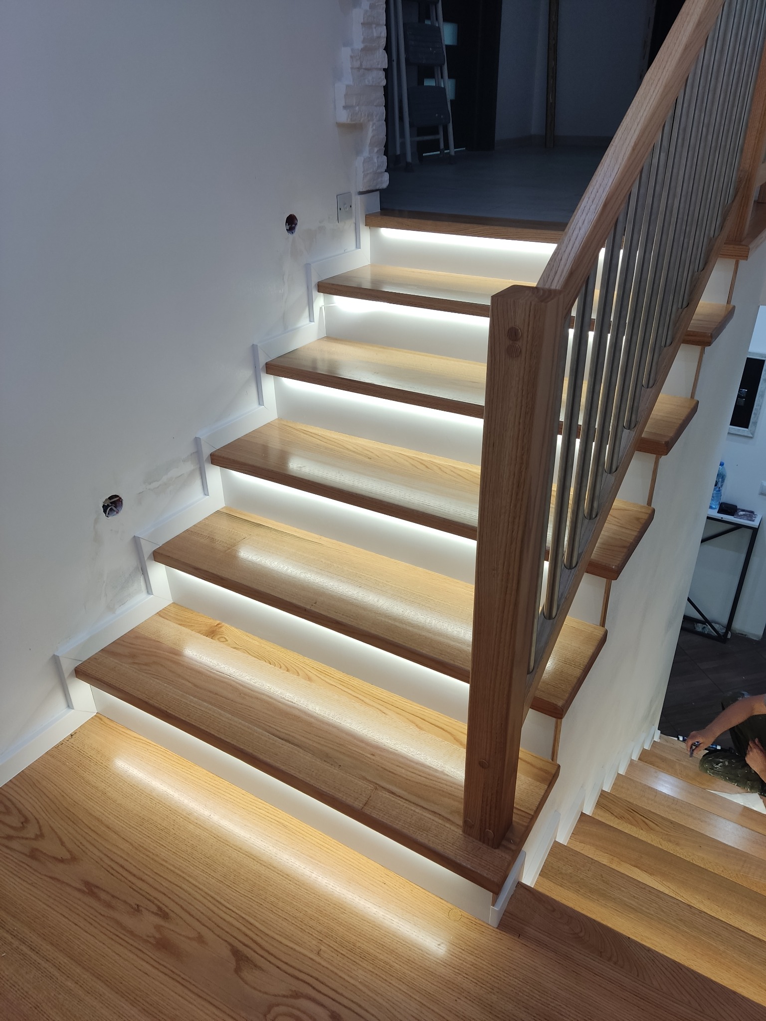 Nowoczesne schody z podświetleniem LED, drewniane stopnie o jasnym odcieniu, metalowa balustrada. Widok z dołu, oświetlenie podkreśla strukturę drewna.
