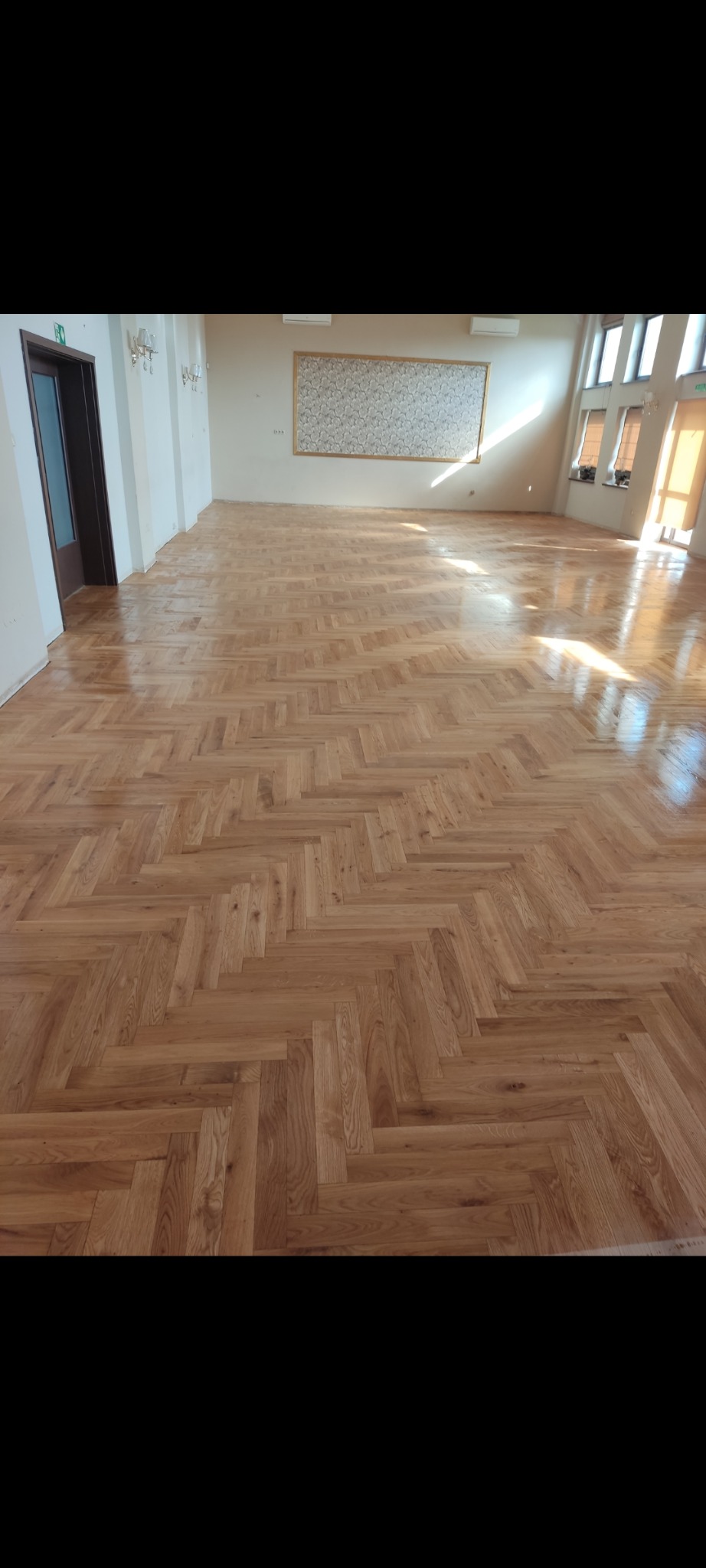 Parkiet ułożony w jodełkę w przestronnym, jasnym wnętrzu. Widoczne naturalne usłojenie drewna i połysk lakieru. W tle okna i drzwi.